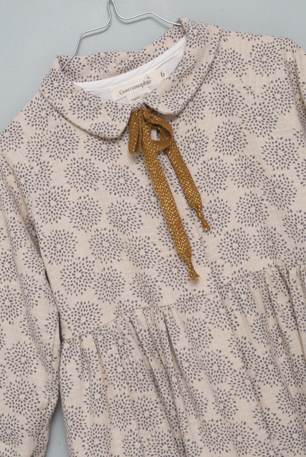 Gemini is an open front dress so you can wear it as a coat. It is made of an ecru jacquard fabric with a gray dahlia print. The buttons are hidden under a placket. You'll love its raglan sleeves and the gold bow detail on the neck.