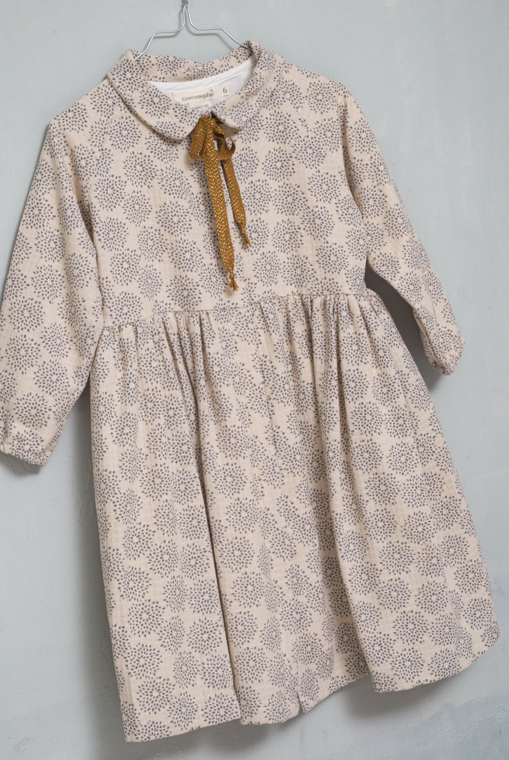 Gemini is an open front dress so you can wear it as a coat. It is made of an ecru jacquard fabric with a gray dahlia print. The buttons are hidden under a placket. You'll love its raglan sleeves and the gold bow detail on the neck.