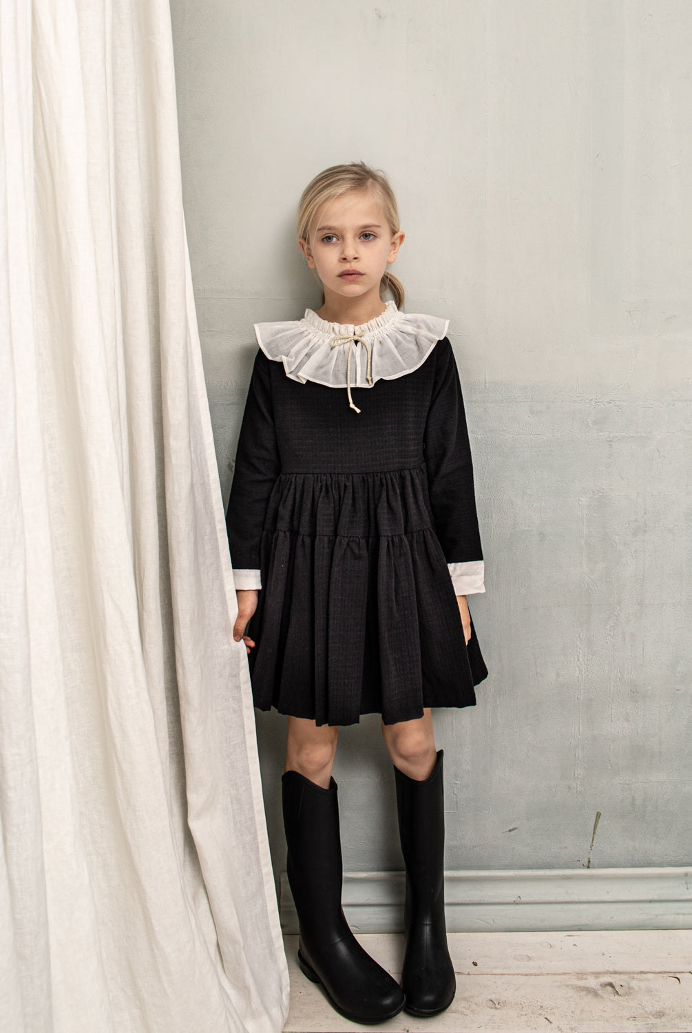 Unos de los vestido más románticos de la colección. Realizado en algodón negro con jacquard en vichy. Te encantará su enorme cuello fruncido en muselina de color blanco roto. Pequeño detalle de lazada en el cuello en tono crudo y dorado. La manga lleva a contraste un detalle de muselina en color blanco roto. Falda con doble volante. Se abotona en la espalda.