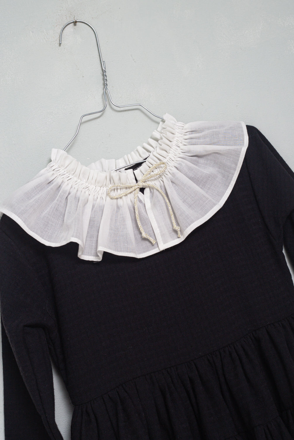 One of the most romantic dresses in the collection. Made of black cotton with gingham jacquard. You'll love its huge, gathered collar in off-white muslin. Small bow detail on the neck in an ecru and gold tone. The sleeve has a contrasting off-white muslin detail. Double flounce skirt. It buttons in the back.