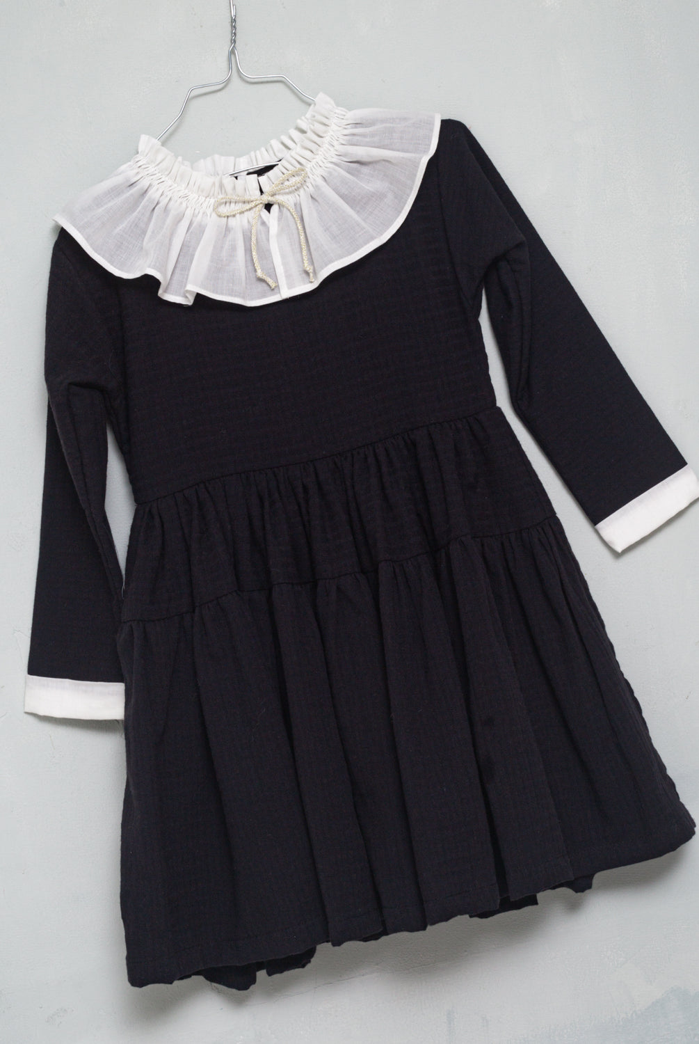 One of the most romantic dresses in the collection. Made of black cotton with gingham jacquard. You'll love its huge, gathered collar in off-white muslin. Small bow detail on the neck in an ecru and gold tone. The sleeve has a contrasting off-white muslin detail. Double flounce skirt. It buttons in the back.