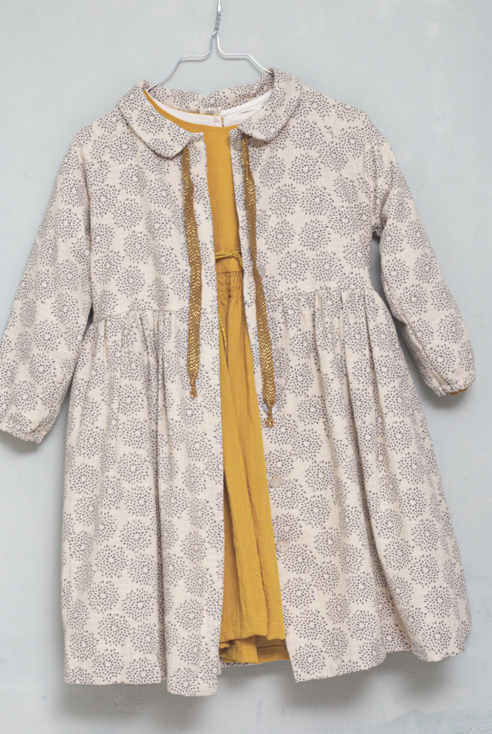 Gemini es un vestido abierto por delante para que puedas usarlo como abrigo. Esta realizado en un tejido de jacquard en color crudo con estampado de dalias en color gris. Los botones estan ocultos bajo una tapeta. Te encantará su manga ranglan y el detalle de lazada dorada en el cuello